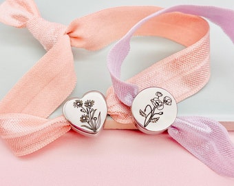May include: Two elastic hair ties with metal charms. One charm is a heart shape with a floral design. The other charm is round with a floral design.