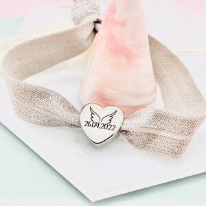 May include: A silver heart charm bracelet with a silver heart charm engraved with a pair of wings and the date 26.04.2022. The bracelet is made of a light gray elastic band.