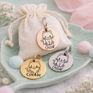 May include: Three round pet tags with cat illustrations and names. One tag is gold-colored with the name "Cookie", another is silver with "Oscar", and the third is rose gold with "Oscar". A small drawstring bag is behind the tags.