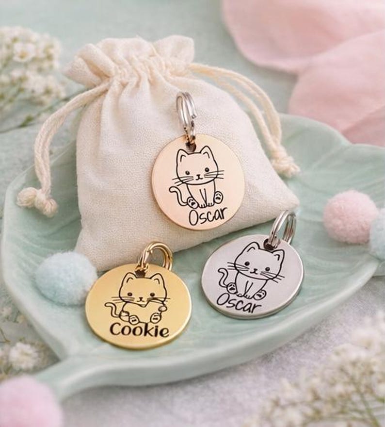 May include: Three round pet tags with cat illustrations and names. One tag is gold-colored with the name "Cookie", another is silver with "Oscar", and the third is rose gold with "Oscar". A small drawstring bag is behind the tags.
