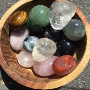 May include: A wooden bowl containing a selection of polished gemstones. The stones include clear quartz, rose quartz, green aventurine, tiger's eye, and other colourful stones. The bowl is made of light brown wood.