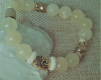 Op de afbeelding: Een witte jade kralen armband met gouden Boeddha-kop accenten. De armband ligt op een lichtbruine stoffen achtergrond.