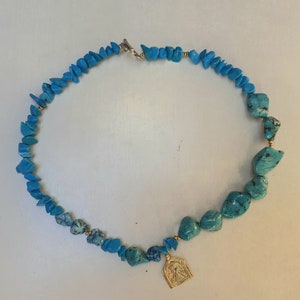 Puede incluir: Un collar de piedras turquesa con un colgante de oro que presenta un icono religioso. El collar está hecho de piedras de forma irregular y está enhebrado en una cadena de oro.