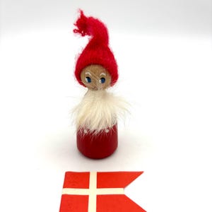May include: A wooden gnome figurine with a red hat and body, white beard, and blue eyes. The figurine is next to a red and white Danish flag. The gnome is a charming decorative item.