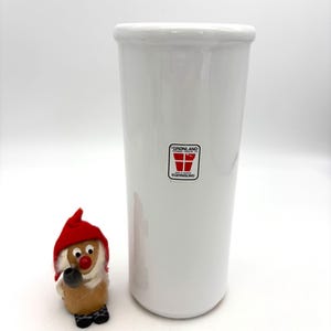 May include: A tall, white ceramic vase with a cylindrical shape. The vase has a small, rectangular label with red and white text. A small gnome figurine with a red hat and pipe is next to the vase.