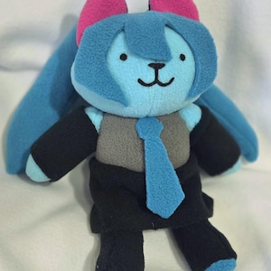 May include: A plush toy in the shape of a cartoon character. The toy has a light blue face and hair, pink ears, and is wearing a gray vest, black suit, and blue tie. The toy has a smiling face.