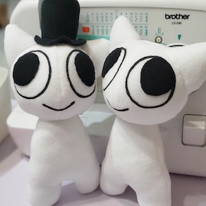 Puede incluir: Dos juguetes de peluche blancos con ojos y bocas negras. Un juguete lleva un sombrero de copa negro. Los juguetes están sentados en una máquina de coser blanca con la marca "brother" y el número de modelo "LS-590" impresos en ella.