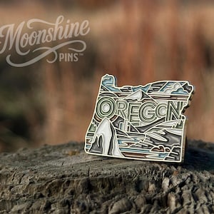 May include: A metal enamel pin shaped like the state of Oregon, featuring a stylized landscape with mountains, trees, and water. The word OREGON is prominently displayed in the center. The pin uses a palette of muted greens, blues, browns, and white.