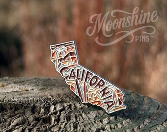 May include: A layered pin in the shape of California, featuring a design of palm trees, mountains, and the word "CALIFORNIA." The pin has a gradient of colors, including white, brown, and orange. The text "Moonshine Pins" is in the upper right corner.