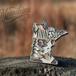May include: A metal enamel pin shaped like the state of Minnesota, featuring the text MINNESOTA in bold letters. The design includes a pine tree, a flower, and geometric landscape patterns in shades of brown, cream, white, and black.