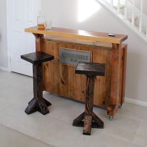 May include: A wooden bar with a metal sign that reads "Armenian Family Bar". The bar has two wooden stools with a dark brown finish. The bar is in a home with a white staircase and a framed picture on the wall.