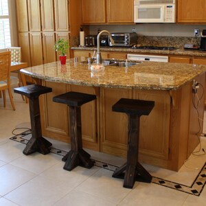 May include: A kitchen island with a granite countertop and three black wooden bar stools. The island has a light wood base and is situated in a kitchen with light wood cabinets and a tiled floor.