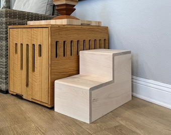 May include: Two-step wooden pet stairs in a light wood finish. The stairs are in a living room setting with a wooden end table and a wicker chair.