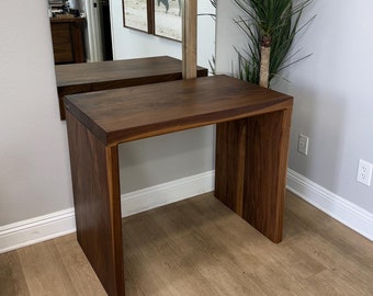 May include: A dark brown wooden desk with a simple design. The desk has a rectangular top and two legs on each side. The desk is sitting on a light brown wood floor.