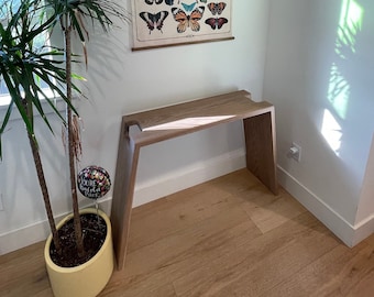 May include: A wooden console table with a unique curved design, featuring a light wood finish. The table is positioned in a corner of a room with a plant and a butterfly poster on the wall.
