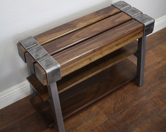 Puede incluir: Un banco de madera con un marco de metal y dos estantes. El banco está hecho de madera oscura y tiene un acabado natural. El marco de metal es plateado y tiene un acabado cepillado.