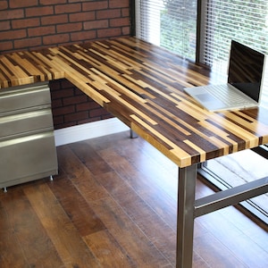 May include: A large, L-shaped desk with a wood top made of various wood types. The desk has a metal frame and a silver filing cabinet.