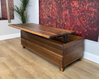 May include: A walnut-finished wooden storage bench with a lift-up lid. The lid is open, showing the interior storage. The bench is supported by short, tapered legs. A large abstract painting in red and brown tones hangs on the wall behind.