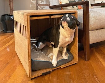 May include: A wooden dog crate with a black and white dog inside. The crate has a slatted design on the front and a grey cushion inside. The crate is sitting on a wooden floor.