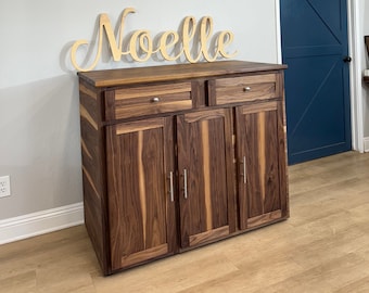 May include: A wooden cabinet with four doors and two drawers. The cabinet is made of walnut wood and has a light brown finish. The cabinet has silver handles. The cabinet is in a room with a white ceiling and white walls. The name "Noelle" is written in cursive on the wall above the cabinet.