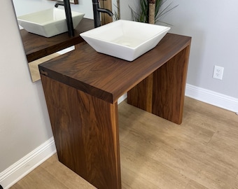 May include: A white ceramic vessel sink sits on a dark brown wooden vanity with a rectangular top and four legs. The vanity is in a bathroom with light brown wood flooring.