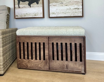 May include: A wooden dog crate with a cushioned top, featuring a slatted design on the front doors. The crate is brown and has a light grey cushion. Two framed landscape prints are hanging on the wall behind the crate.