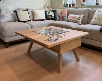 May include: A light oak square coffee table with angled legs. A board game is set up on the table. The table is in a living room with a grey sectional sofa and decorative cushions. A hanging plant is visible.