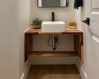 Puede incluir: Un lavabo de baño de porcelana blanca con un grifo negro se asienta sobre una encimera de madera con un estante. La encimera está hecha de madera recuperada con una variedad de tonos de madera. Una pequeña planta en maceta se encuentra sobre la encimera a la izquierda del lavabo.