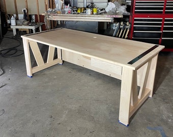 May include: A light wood desk with a simple design. The desk has a rectangular top and two long legs on each side. The legs are angled inwards and meet at the bottom. The desk has a drawer on the front side. The desk is in a workshop setting.