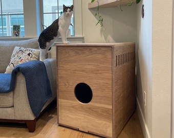 May include: A light wood cat litter box with a round opening and a slatted vent on the side. A cat is sitting on top of the box.