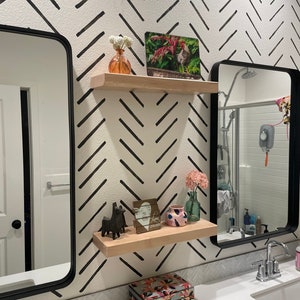 Könnte beinhalten: Ein Badezimmer mit einer weißen und schwarzen Chevron-Musterwand. Zwei schwarze Spiegel hängen über einer weißen Arbeitsplatte mit einem weißen Waschbecken. Zwei Holzregale mit Dekoartikeln, darunter eine Vase mit Blumen, ein Holzschild mit dem Text "Love Lives Here" und ein kleiner Keramik-Lama, sind an der Wand montiert.