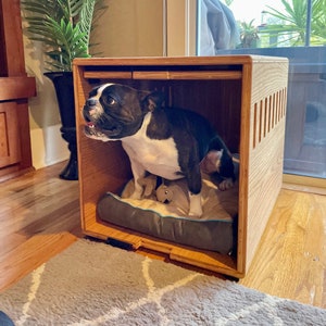 May include: A black and white Boston Terrier dog standing in a wooden dog crate with a gray bed and a stuffed toy. The crate has a slatted front and a wooden top.