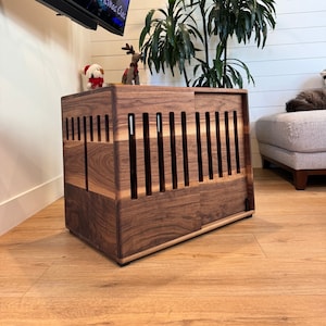 May include: A wooden dog crate with a sliding door. The crate is made of walnut wood and has a slatted design. The crate is sitting on a wooden floor.
