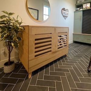 May include: A light wood cabinet with two doors and a gold-framed round mirror above it. The cabinet has a slatted design on the doors and sits on four legs. The cabinet is in a room with a green tiled floor and a green cabinet in the background.