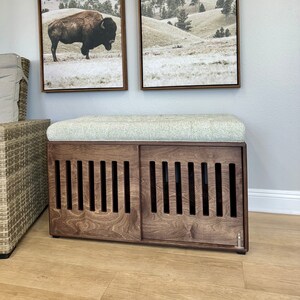 May include: A wooden dog crate with a cushioned top, featuring a slatted design on the front doors. The crate is brown and has a light gray cushion. Two framed landscape prints are hanging on the wall behind the crate.