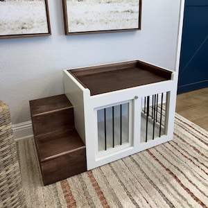 May include: White wooden dog crate with a brown wooden top and steps. The crate has a metal door with vertical bars. The crate is sitting on a striped rug.
