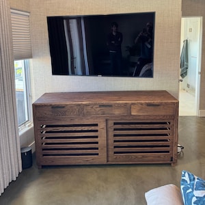May include: A dark brown wooden media console with three drawers and two slatted doors. The console is in front of a wall-mounted television.