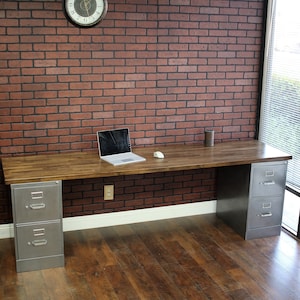 May include: A wooden desk with two metal file cabinets. The desk has a light wood finish and the file cabinets are silver. The desk is in front of a brick wall and a window.
