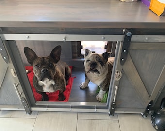 May include: Two dogs, a brindle French Bulldog and a white English Bulldog, are inside a wooden dog crate with a grey painted finish. The crate has a metal door with a handle and a metal latch. The dogs are looking at the camera.