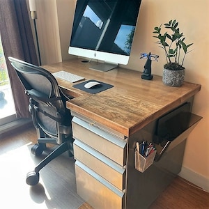 May include: A wooden desk with a metal filing cabinet and a computer monitor. The desk has a light wood finish and the filing cabinet has a brushed metal finish. There is a black office chair with a mesh back and a potted plant on the desk.