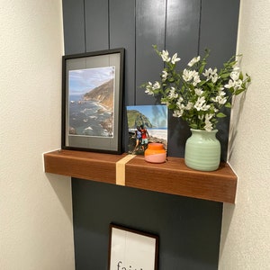 Könnte beinhalten: Ein Holzregal mit zwei gerahmten Bildern und einer Vase mit weißen Blumen. Das Regal ist an einer dunkelgrauen Wand montiert. Die Bilder zeigen eine Strandszene und ein Paar, das am Strand steht. Die Vase ist hellgrün und hat eine strukturierte Oberfläche. Der Text "faith" ist auf weißem Hintergrund in einem schwarzen Rahmen gedruckt.
