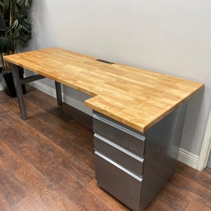 May include: A large, L-shaped desk with a light wood top and a metal base. The desk has a built-in power strip and a set of three drawers on the right side.