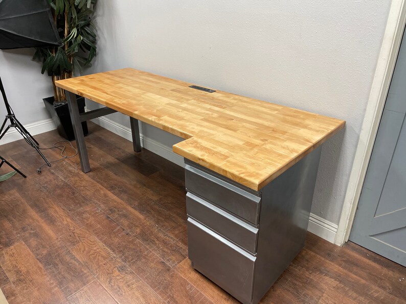May include: A large, L-shaped desk with a light wood top and a metal base. The desk has a built-in power strip and a set of three drawers on the right side.