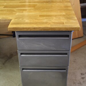 May include: A wooden desk with a gray metal drawer unit. The desk top is made of light wood planks and the drawers are a dark gray color.