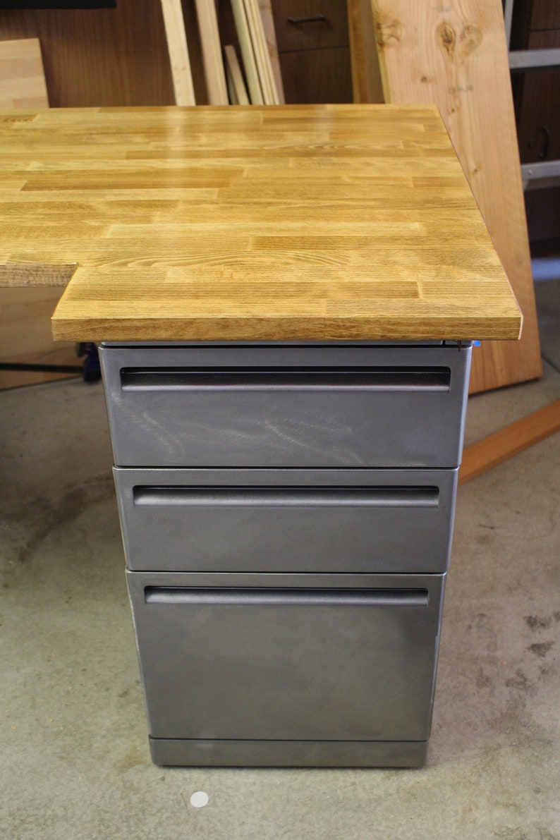 May include: A wooden desk with a gray metal drawer unit. The desk top is made of light wood planks and the drawers are a dark gray color.