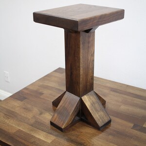 May include: A dark brown wooden end table with a square top and a pedestal base. The base has a unique, geometric design with four legs that extend out from the center.