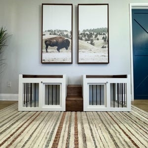 May include: Two white dog crates with black metal bars and a wooden step in between. The crates are set against a white wall with two framed landscape prints above them. The floor is covered in a striped rug.