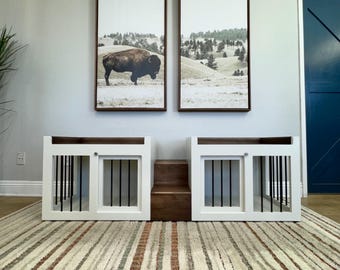 May include: Two white dog crates with black metal bars and a wooden step in between. The crates are set against a white wall with two framed landscape prints above them. The floor is covered in a striped rug.