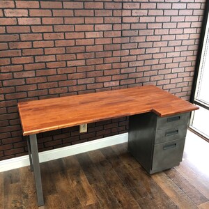 May include: A wooden desk with a metal frame and a metal filing cabinet. The desk has a light wood top and the filing cabinet has a gray finish. The desk is in front of a brick wall.
