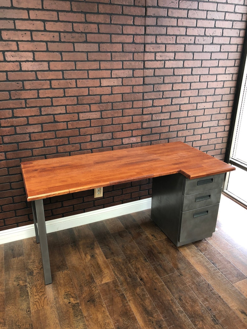May include: A wooden desk with a metal frame and a metal filing cabinet. The desk has a light wood top and the filing cabinet has a gray finish. The desk is in front of a brick wall.